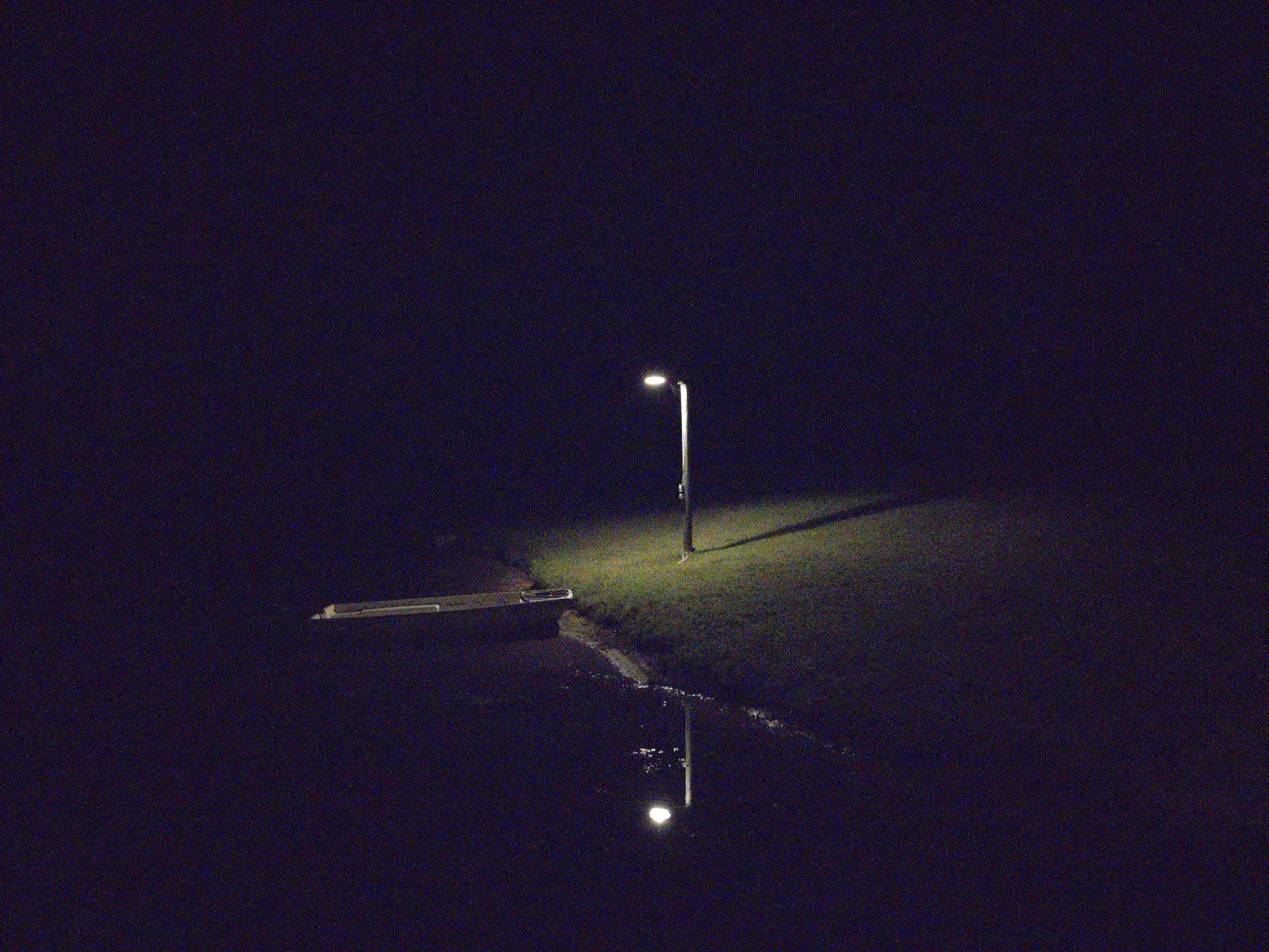 A photograph taken at night of a small boat grounded halfway onto the shore of a lake, with an incandescent light illuminating it from above.