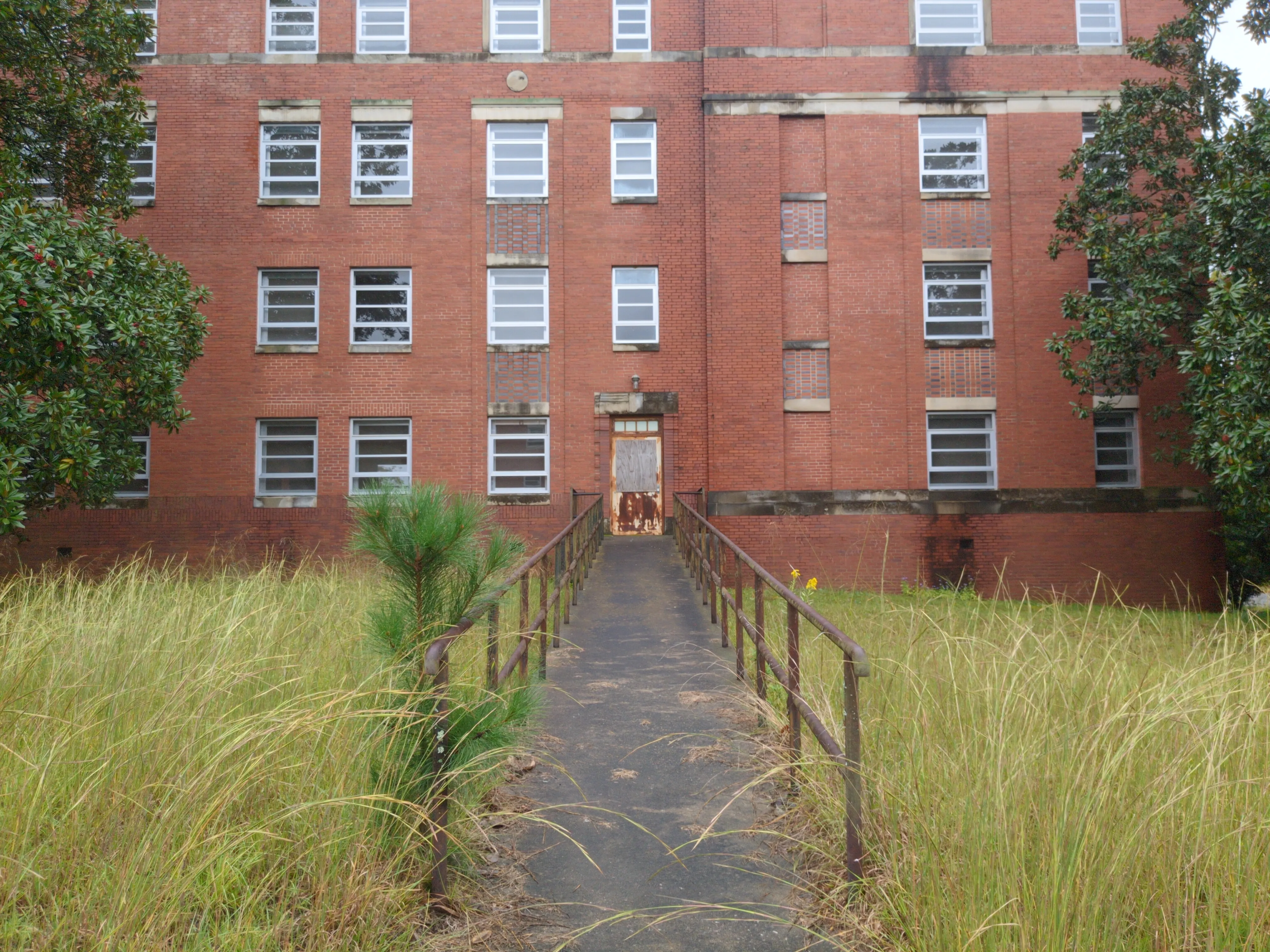 A photograph of a gently-inclined ramp leading to a rusty door. The grass on each side of the ramp is overgrown.